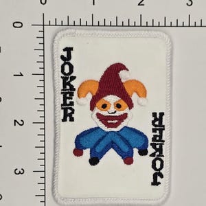 Op de afbeelding: Een witte geborduurde patch met een rood, oranje en blauw jokerkaartontwerp. De tekst "JOKER" is aan beide kanten van de kaart geborduurd.