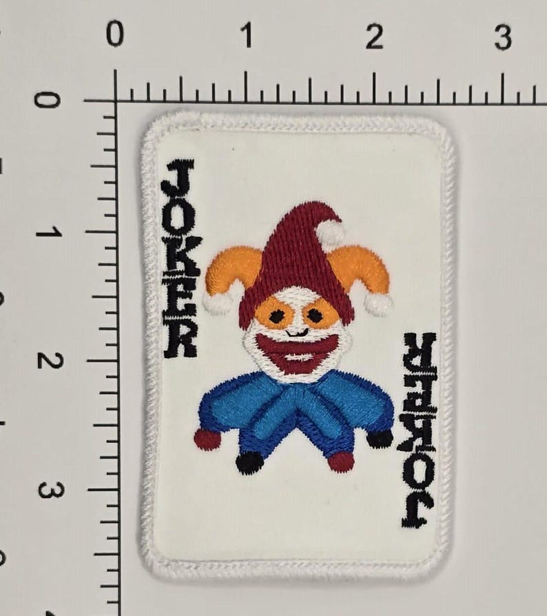 Op de afbeelding: Een witte geborduurde patch met een rood, oranje en blauw jokerkaartontwerp. De tekst "JOKER" is aan beide kanten van de kaart geborduurd.