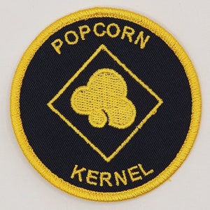May include: A black and yellow embroidered patch with the words "Popcorn Kernel" and a yellow popcorn kernel design inside a yellow diamond.