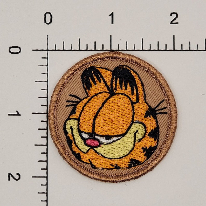 May include: Round embroidered patch featuring Garfield the cat's face. The patch has a tan background with a brown border. Garfield's face is orange with black markings, closed eyes, and a pink tongue.