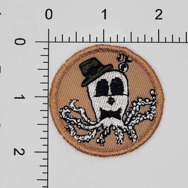 Puede incluir: Un parche bordado marr&oacute;n con un pulpo blanco que lleva un sombrero verde y una pajarita negra. El pulpo tiene ojos negros y una cara blanca.