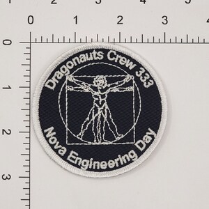 Op de afbeelding: Een geborduurde zwarte en witte patch met een witte cirkel rond de rand. Het midden van de patch heeft een witte omtrek van een menselijke figuur in een cirkel en een vierkant. De tekst "Dragonaunts Crew 333 Nova Engineering Day" staat in een witte boog rond de figuur.