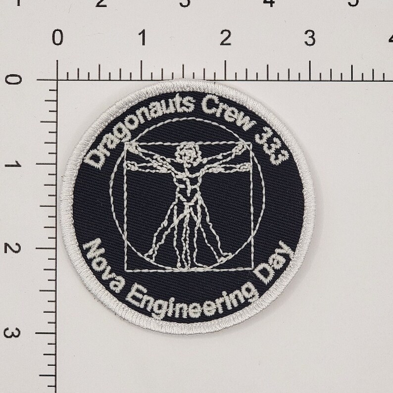 Op de afbeelding: Een geborduurde zwarte en witte patch met een witte cirkel rond de rand. Het midden van de patch heeft een witte omtrek van een menselijke figuur in een cirkel en een vierkant. De tekst "Dragonaunts Crew 333 Nova Engineering Day" staat in een witte boog rond de figuur.