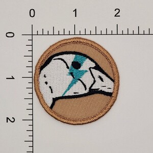 May include: A round embroidered patch with a tan background and a white bird with a blue lightning bolt design. The bird is facing left.