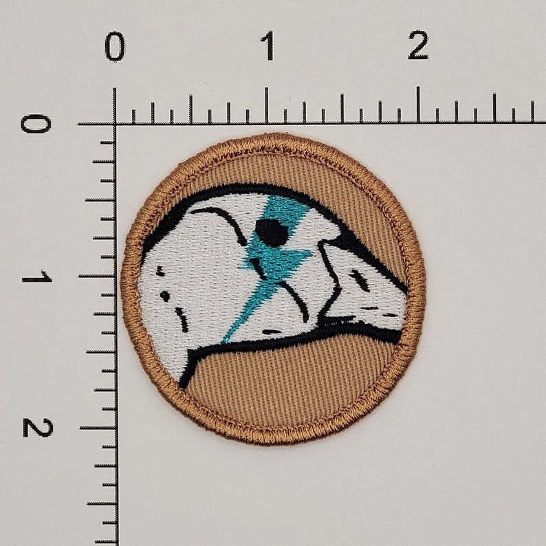 May include: A round embroidered patch with a tan background and a white bird with a blue lightning bolt design. The bird is facing left.