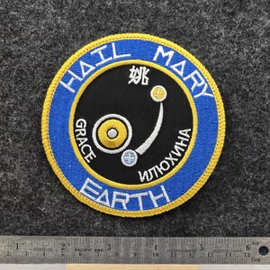 Op de afbeelding: Geborduurde patch met een cirkelvormig ontwerp met de woorden "HAIL MARY EARTH" in een blauwe en gele rand. Het midden heeft een zwarte achtergrond met witte en gele afbeeldingen en tekst. De patch bevindt zich op een grijze vilt achtergrond.