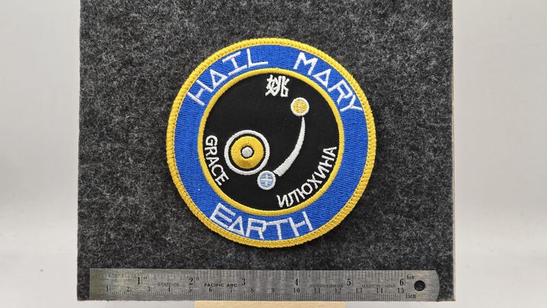 Op de afbeelding: Geborduurde patch met een cirkelvormig ontwerp met de woorden "HAIL MARY EARTH" in een blauwe en gele rand. Het midden heeft een zwarte achtergrond met witte en gele afbeeldingen en tekst. De patch bevindt zich op een grijze vilt achtergrond.