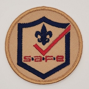 Puede incluir: Un parche redondo de color beige con un escudo azul y blanco con una marca de verificaci&oacute;n roja y la palabra "SAFE" en letras rojas.