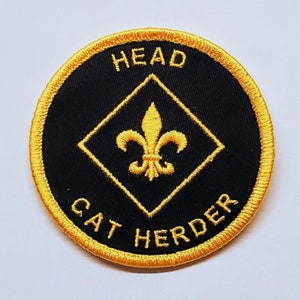 May include: Round black patch with gold embroidered text and design. The text reads "HEAD" at the top and "CAT HERDER" at the bottom. A gold fleur-de-lis is centered inside a gold diamond.