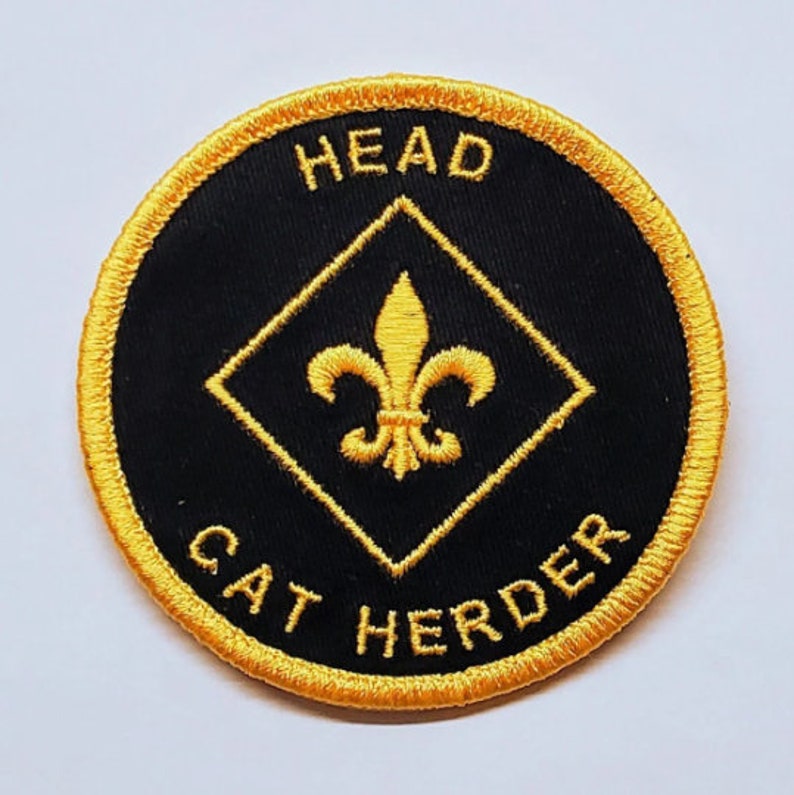 May include: Round black patch with gold embroidered text and design. The text reads "HEAD" at the top and "CAT HERDER" at the bottom. A gold fleur-de-lis is centered inside a gold diamond.