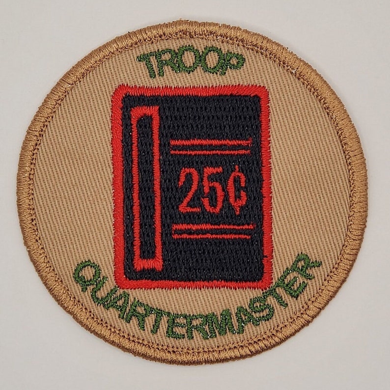 Pu&ograve; includere: Una toppa circolare color beige con testo ricamato in verde che recita "TROOP QUARTERMASTER" e un'immagine ricamata in nero e rosso di un libro mastro con il testo "25&cent;" all'interno.