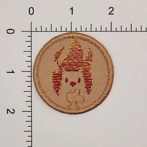 May include: Round embroidered patch with a tan background and a red and yellow cartoon-like design. The design features a stylized creature with a head, ears, and a small body. The patch has a stitched border.
