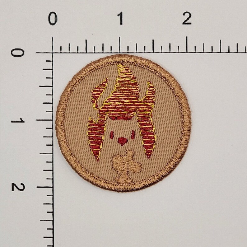 May include: Round embroidered patch with a tan background and a red and yellow cartoon-like design. The design features a stylized creature with a head, ears, and a small body. The patch has a stitched border.
