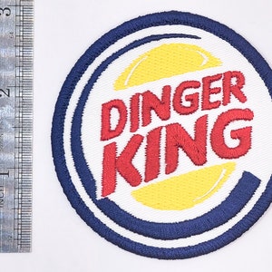 K&ouml;nnte beinhalten: Ein wei&szlig;er, blauer und gelber gestickter Aufn&auml;her mit dem Text "DINGER KING" in roten Buchstaben. Der Aufn&auml;her ist rund und hat einen blauen Rand.