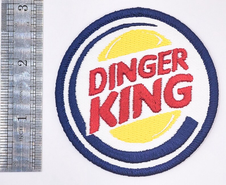 K&ouml;nnte beinhalten: Ein wei&szlig;er, blauer und gelber gestickter Aufn&auml;her mit dem Text "DINGER KING" in roten Buchstaben. Der Aufn&auml;her ist rund und hat einen blauen Rand.