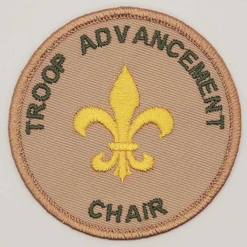 May include: A tan round patch with green embroidered text that reads "Troop Advancement Chair" and a yellow embroidered fleur-de-lis in the center.