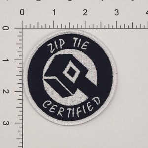 Może przedstawiać: Naszywka haftowana w kolorze białym i czarnym z tekstem "Zip Tie Certified" w k&oacute;łku.