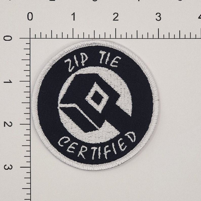Może przedstawiać: Naszywka haftowana w kolorze białym i czarnym z tekstem "Zip Tie Certified" w k&oacute;łku.