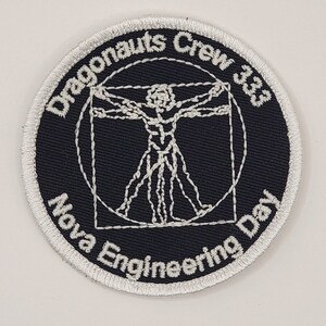 Op de afbeelding: Een geborduurde zwarte en witte patch met een wit Vitruvius-manontwerp in het midden. De tekst "Dragonaunts Crew 3.03 Nova Engineering Day" is rond de buitenkant van de patch geborduurd.