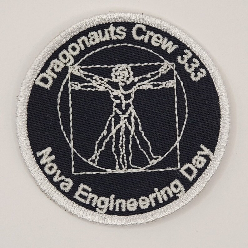 Op de afbeelding: Een geborduurde zwarte en witte patch met een wit Vitruvius-manontwerp in het midden. De tekst "Dragonaunts Crew 3.03 Nova Engineering Day" is rond de buitenkant van de patch geborduurd.