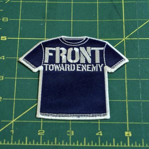 May include: A navy blue patch in the shape of a t-shirt with the words "FRONT TOWARD ENEMY" embroidered in silver thread. The patch has a white border and is set against a green grid background.