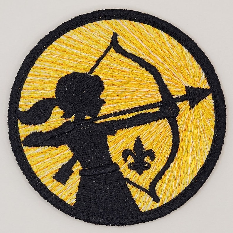 May include: A yellow embroidered patch with a silhouette of a person holding a bow and arrow. The person is wearing a skirt and has long hair. A fleur-de-lis is in the background.
