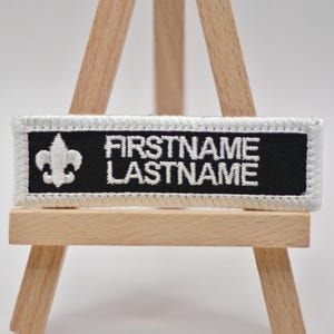 May include: A rectangular black and white embroidered patch with the text "FIRSTNAME LASTNAME" in white. A white fleur-de-lis is on the left side. The patch has a white border.