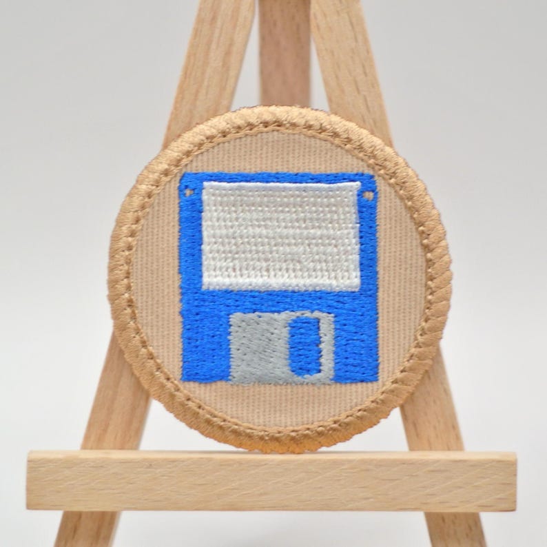 May include: Embroidered patch featuring a blue and gray floppy disk design on a tan background. The patch has a tan border and is displayed on a small wooden easel. The design is a nostalgic nod to vintage technology.
