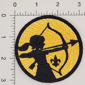 May include: A yellow and white embroidered patch with a black silhouette of a person holding a bow and arrow. The person is wearing a skirt and has long hair. The patch has a yellow fleur-de-lis in the lower right corner.