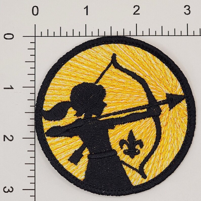 May include: A yellow and white embroidered patch with a black silhouette of a person holding a bow and arrow. The person is wearing a skirt and has long hair. The patch has a yellow fleur-de-lis in the lower right corner.