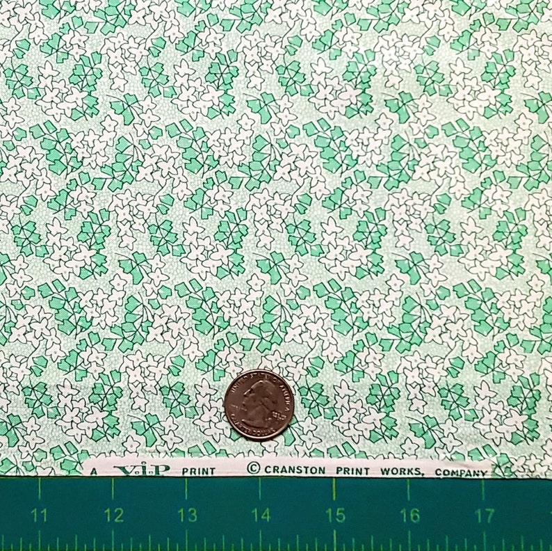Pu&ograve; includere: Un primo piano di un tessuto verde e bianco con un motivo ripetitivo di piccoli fiori bianchi e foglie verdi. Il tessuto &egrave; testurizzato e ha un aspetto vintage. Una moneta da 25 centesimi di dollaro americano &egrave; inclusa per la scala.
