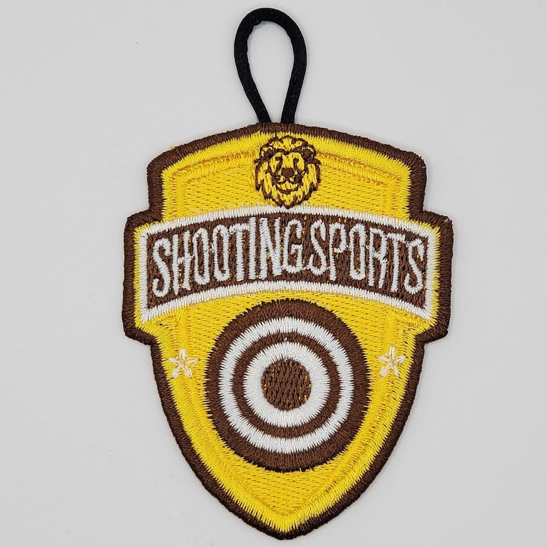 Pu&ograve; includere: Toppa ricamata gialla e marrone con una testa di leone e il testo "Shooting Sports" e un design a bersaglio.