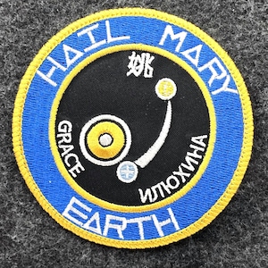 May include: Circular patch with a blue border and yellow trim. The patch features the words "HAIL MARY EARTH" in white, with additional text in Chinese and Cyrillic. A black circle in the center contains a stylized design.