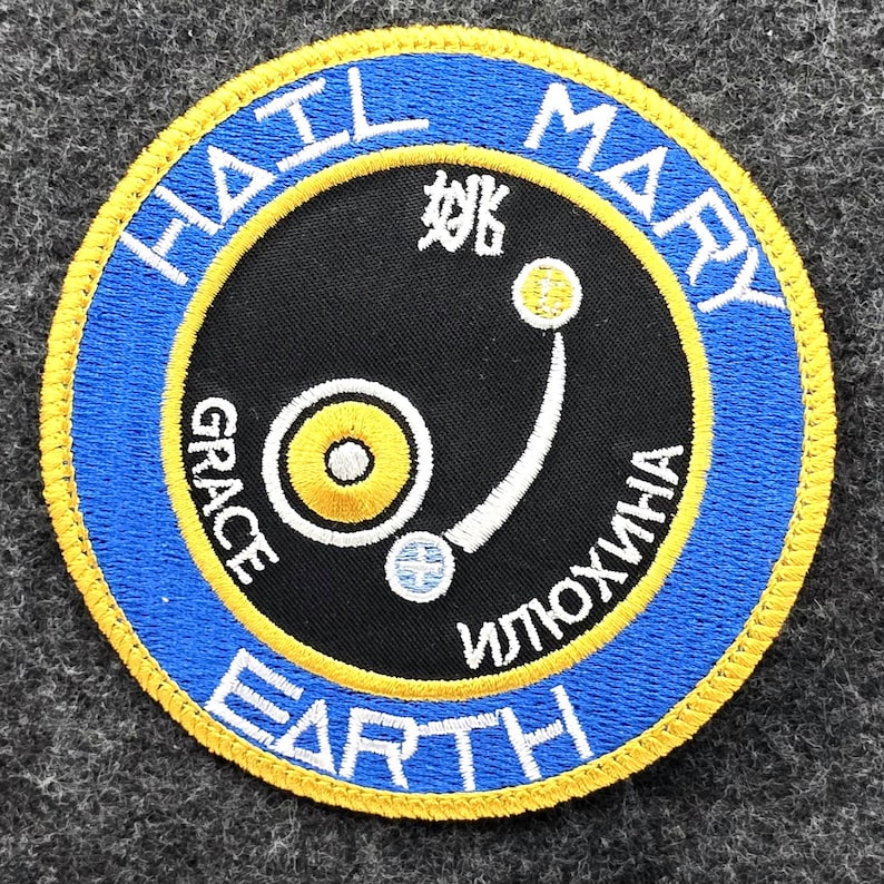 Op de afbeelding: Ronde patch met een blauwe rand en gele rand. De patch bevat de woorden "HAIL MARY EARTH" in het wit, met extra tekst in het Chinees en Cyrillisch. Een zwarte cirkel in het midden bevat een gestileerd ontwerp.