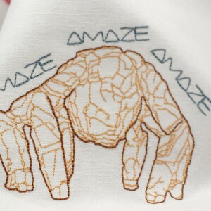 Pu&ograve; includere: Tessuto bianco ricamato con un disegno di creatura marrone e beige. La parola "AMAZE" &egrave; ricamata in verde acqua intorno alla creatura. Il tessuto ha un bordo a righe rosse e bianche.