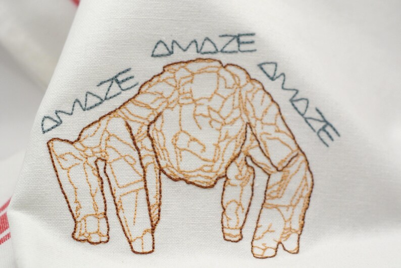 Pu&ograve; includere: Tessuto bianco ricamato con un disegno di creatura marrone e beige. La parola "AMAZE" &egrave; ricamata in verde acqua intorno alla creatura. Il tessuto ha un bordo a righe rosse e bianche.
