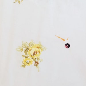 May include: White fabric with a yellow floral pattern. The fabric has a small, dark red button in the center.