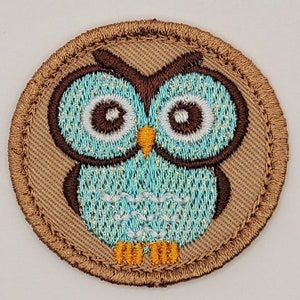 May include: Embroidered patch featuring a cartoon owl. The owl has large, round eyes, a turquoise body, and brown and gold accents. The patch is circular with a brown border and is likely designed to be sewn onto clothing or accessories.