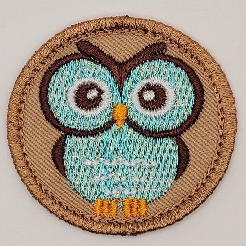 May include: Embroidered patch featuring a cartoon owl. The owl has large, round eyes, a turquoise body, and brown and gold accents. The patch is circular with a brown border and is likely designed to be sewn onto clothing or accessories.