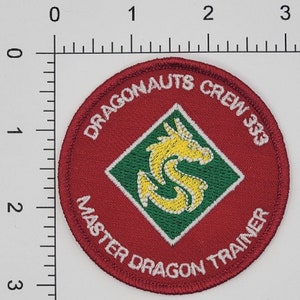 Peut inclure: &Eacute;cusson brod&eacute; rouge avec du texte blanc qui dit "Dragonaunts Crew 383 Master Dragon Trainer". Un diamant vert et blanc avec un dragon jaune est au centre.