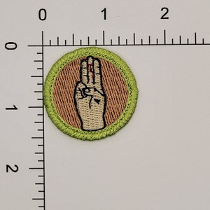 May include: A green and brown embroidered scout patch with a hand making the scout sign.