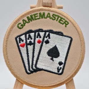 May include: A circular, beige patch embroidered with the word "GAMEMASTER" in green. The patch shows three playing cards, each an Ace. The Ace of spades is visible, along with the Ace of hearts and diamonds.
