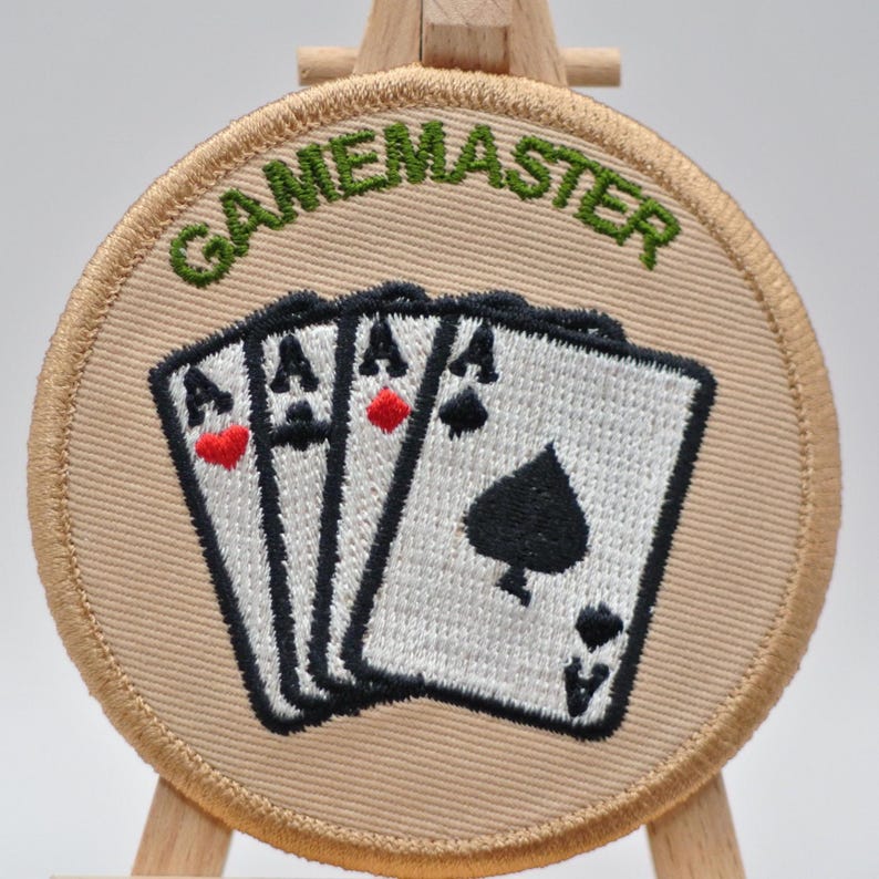 May include: A circular, beige patch embroidered with the word "GAMEMASTER" in green. The patch shows three playing cards, each an Ace. The Ace of spades is visible, along with the Ace of hearts and diamonds.