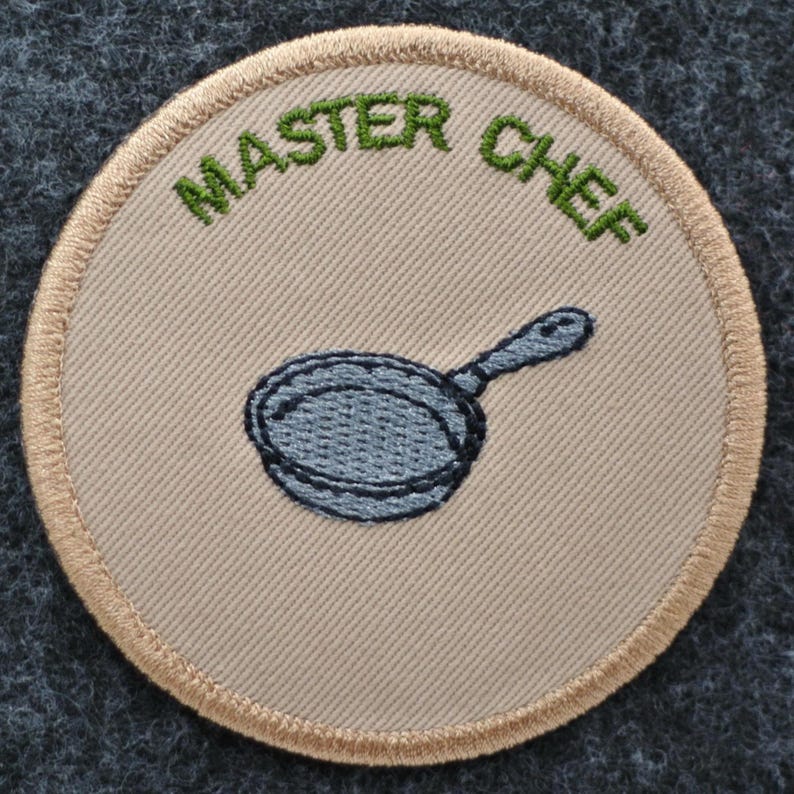 May include: Circular embroidered patch featuring the words "MASTER CHEF" in green above a blue frying pan. The patch has a tan centre and a tan border, set against a dark grey background.
