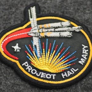 Puede incluir: Parche bordado con fondo negro, con una nave espacial y el texto "PROJECT HAIL MARY". El dise&ntilde;o incluye un arco rojo y amarillo, una estrella y rayos coloridos que emanan de la parte inferior. El parche est&aacute; bordeado de amarillo.