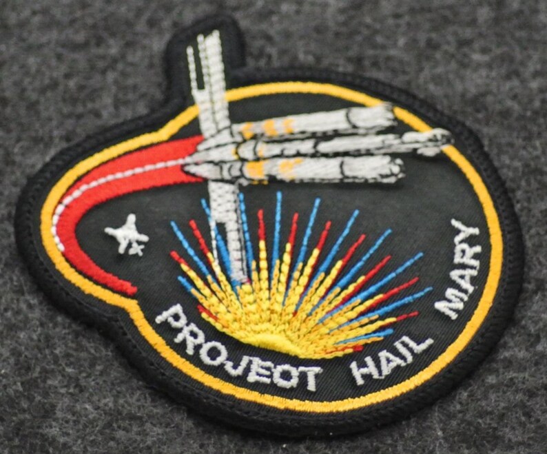 Puede incluir: Parche bordado con fondo negro, con una nave espacial y el texto "PROJECT HAIL MARY". El dise&ntilde;o incluye un arco rojo y amarillo, una estrella y rayos coloridos que emanan de la parte inferior. El parche est&aacute; bordeado de amarillo.