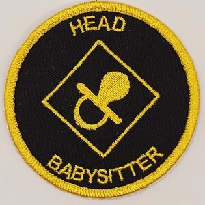 May include: A black and gold embroidered patch with a diamond shape in the center. The diamond shape contains a yellow pacifier. The text "HEAD BABYSITTER" is written around the outside of the patch.