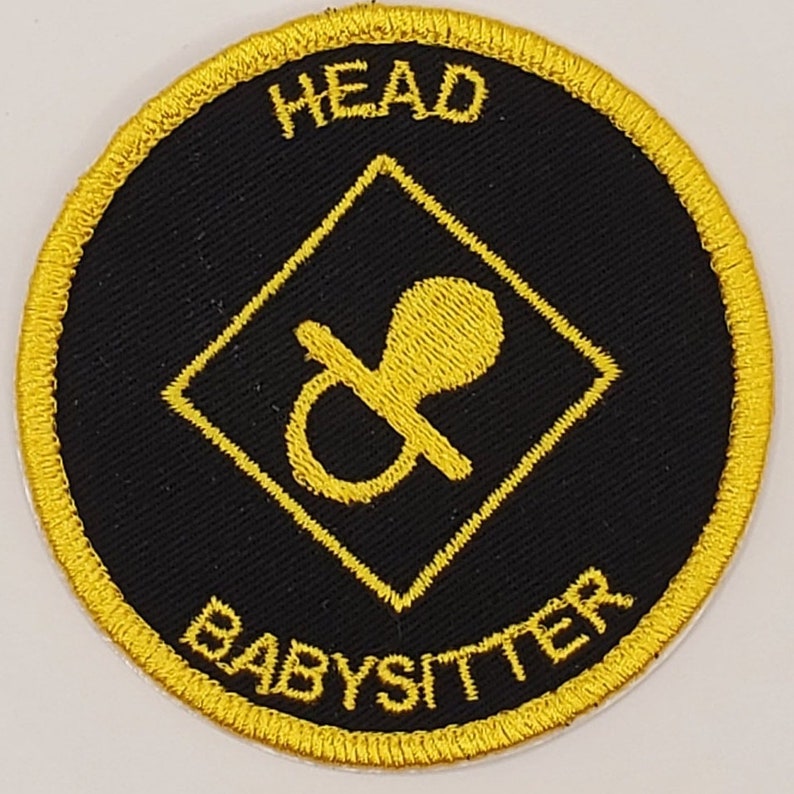 May include: A black and gold embroidered patch with a diamond shape in the center. The diamond shape contains a yellow pacifier. The text "HEAD BABYSITTER" is written around the outside of the patch.