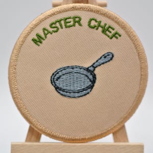 May include: A circular, beige embroidered patch featuring the words "MASTER CHEF" in green above a grey frying pan. The patch has a tan border and is displayed on a wooden easel.