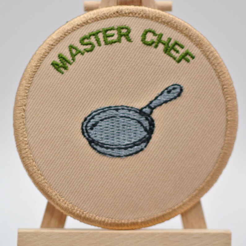 May include: A circular, beige embroidered patch featuring the words "MASTER CHEF" in green above a grey frying pan. The patch has a tan border and is displayed on a wooden easel.
